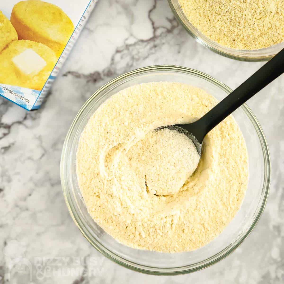 Overhead shot of dry cornbread mix in a clear bowl with a spoon on a marble surface and a box of cornbread mix on the side.