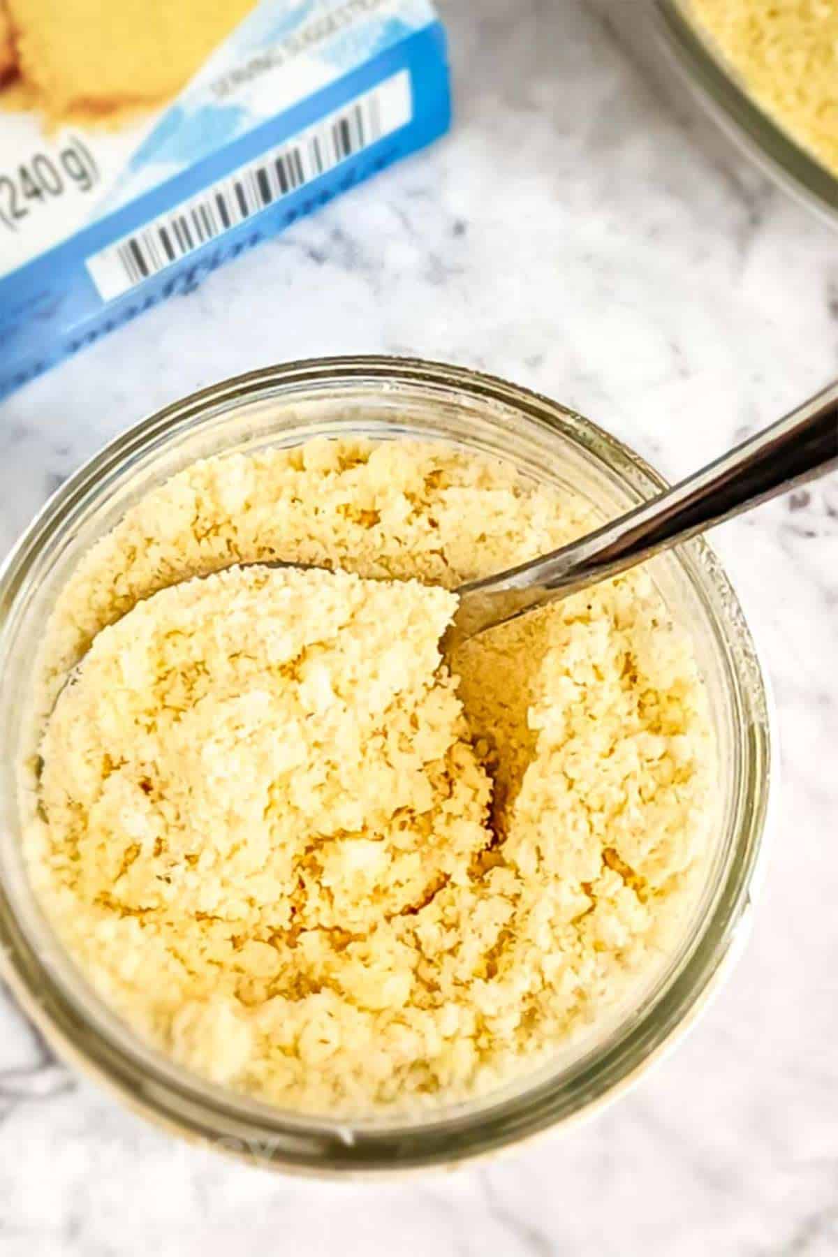 Overhead shot of a small clear bowl with cornbread mix and a spoon on a marble surface and a cornbread mix box in the background.