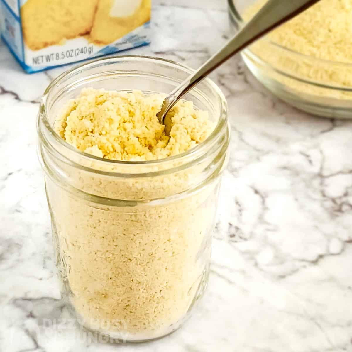 Side shot of cornbread mix in a clear mason jar with a spoon on a marble surface and a box of cornbread mix in the background.