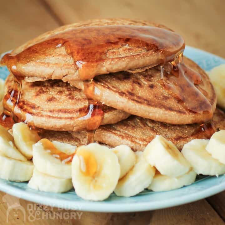 Close up side shot of multiple pancakes stacked on each other with syrup drizzled on them and bananas on the side all on a light blue plate.