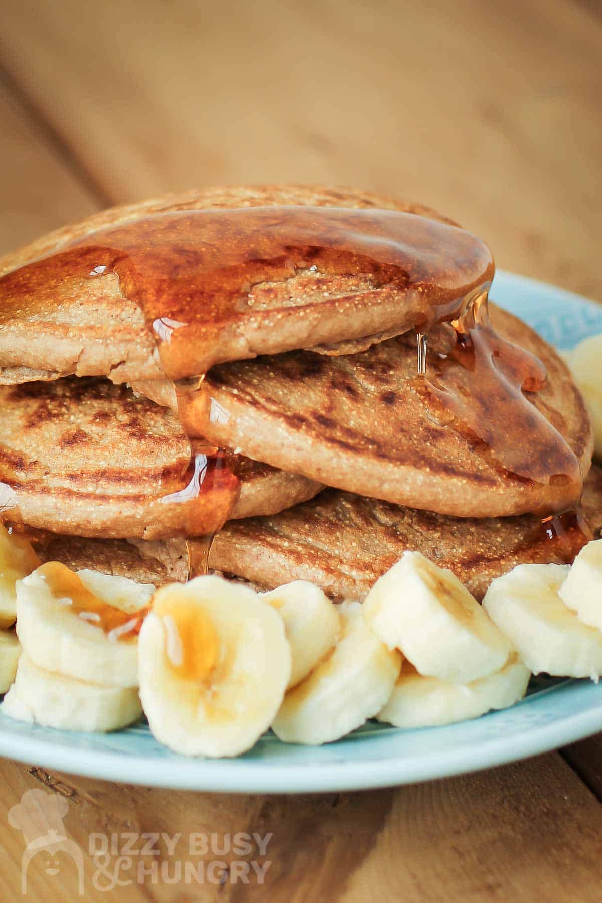 Close up side shot of multiple pancakes stacked on each other with syrup drizzled on them and bananas on the side all on a light blue plate.