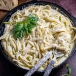 Overhead shot of pesto pasta garnished with basil in a saucepan with silverware on the side and bread in the background.