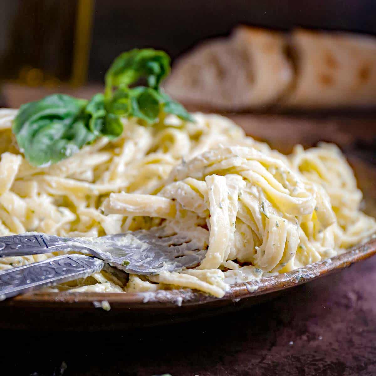 Side shot of pesto pasta garnished with basil in a wooden bowl with silverware on the side and sliced bread in the background.