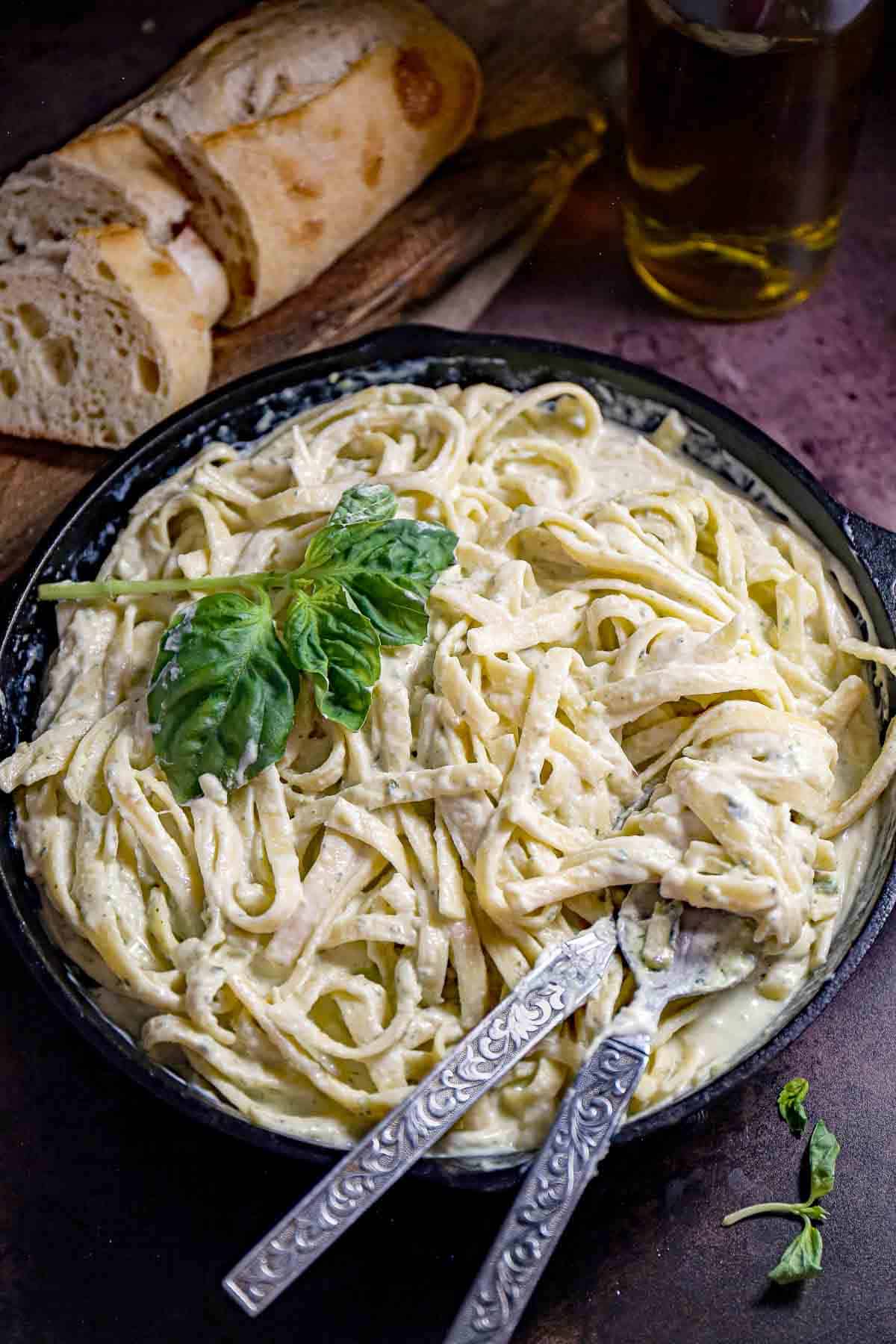 Overhead shot of pesto pasta garnished with basil in a saucepan with silverware on the side and bread in the background.