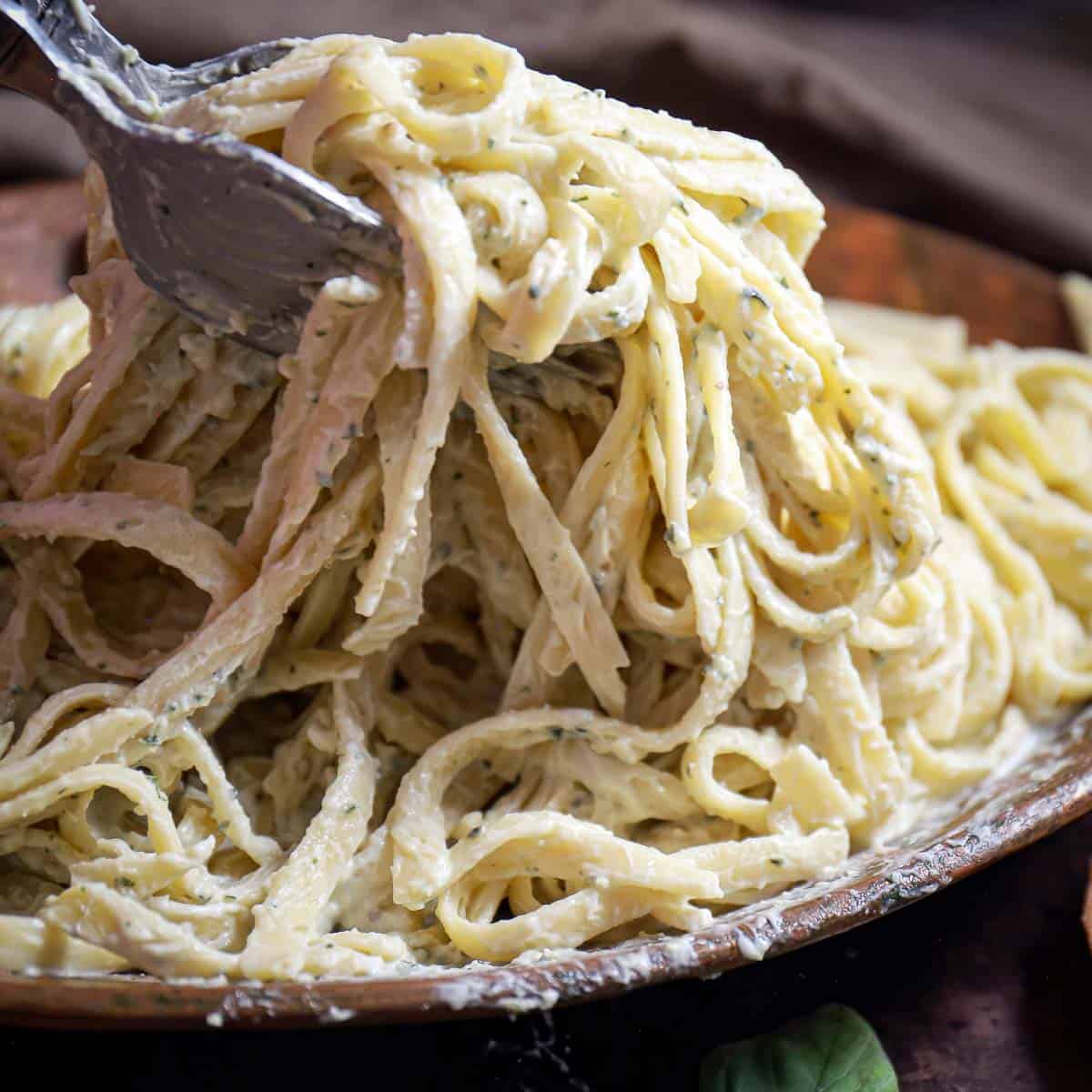 Side close up shot of a fork scooping some pesto pasta off of a brown bowl with basil on the side.