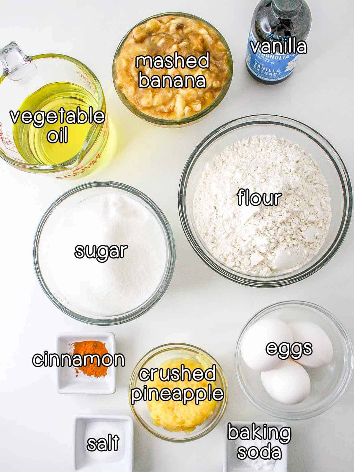 Overhead shot of ingredients - vanilla, mashed banana, vegetable oil, sugar, flour, cinnamon, crushed pineapple, eggs, salt, and baking soda.