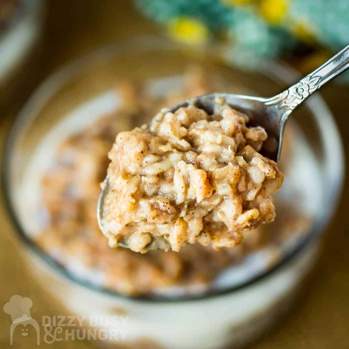 Close up shot of a spoon holding a bite of oatmeal over a clear bowl of oatmeal on a wooden surface.