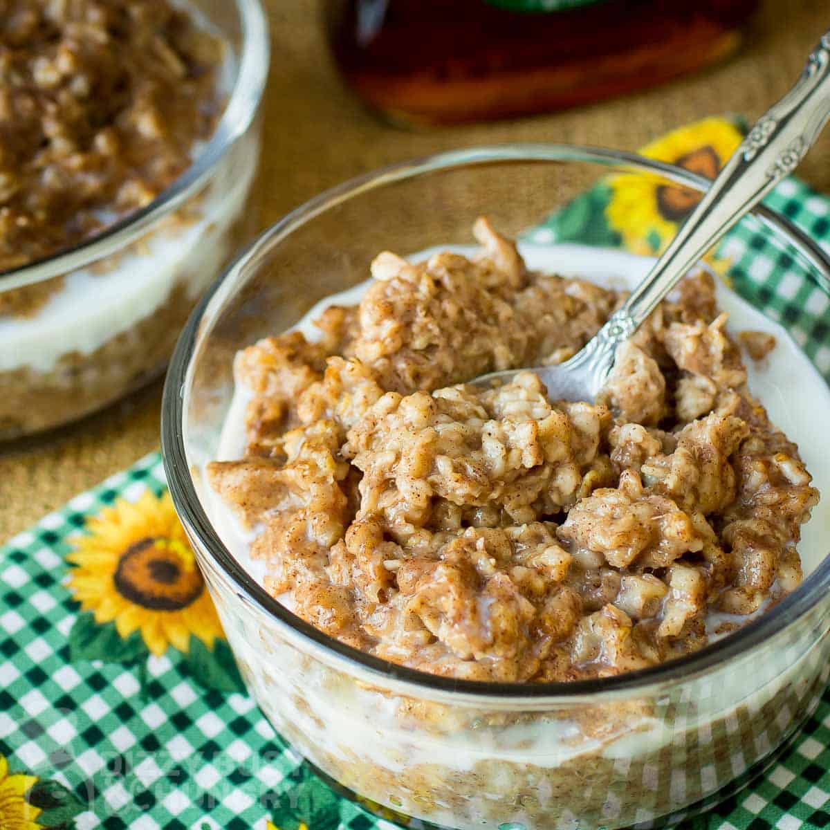 Side angled shot of oatmeal in a clear bowl with a spoon on a green checkered cloth with sunflowers.