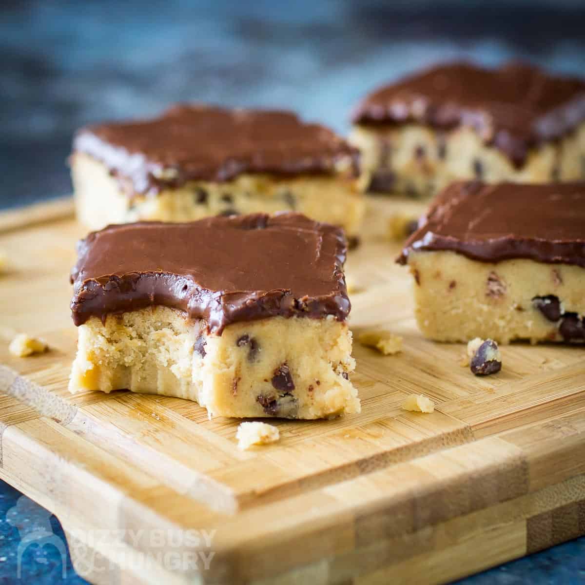 Side shot of cookie dough bars with chocolate glaze on a wooden cutting board on a blue surface.