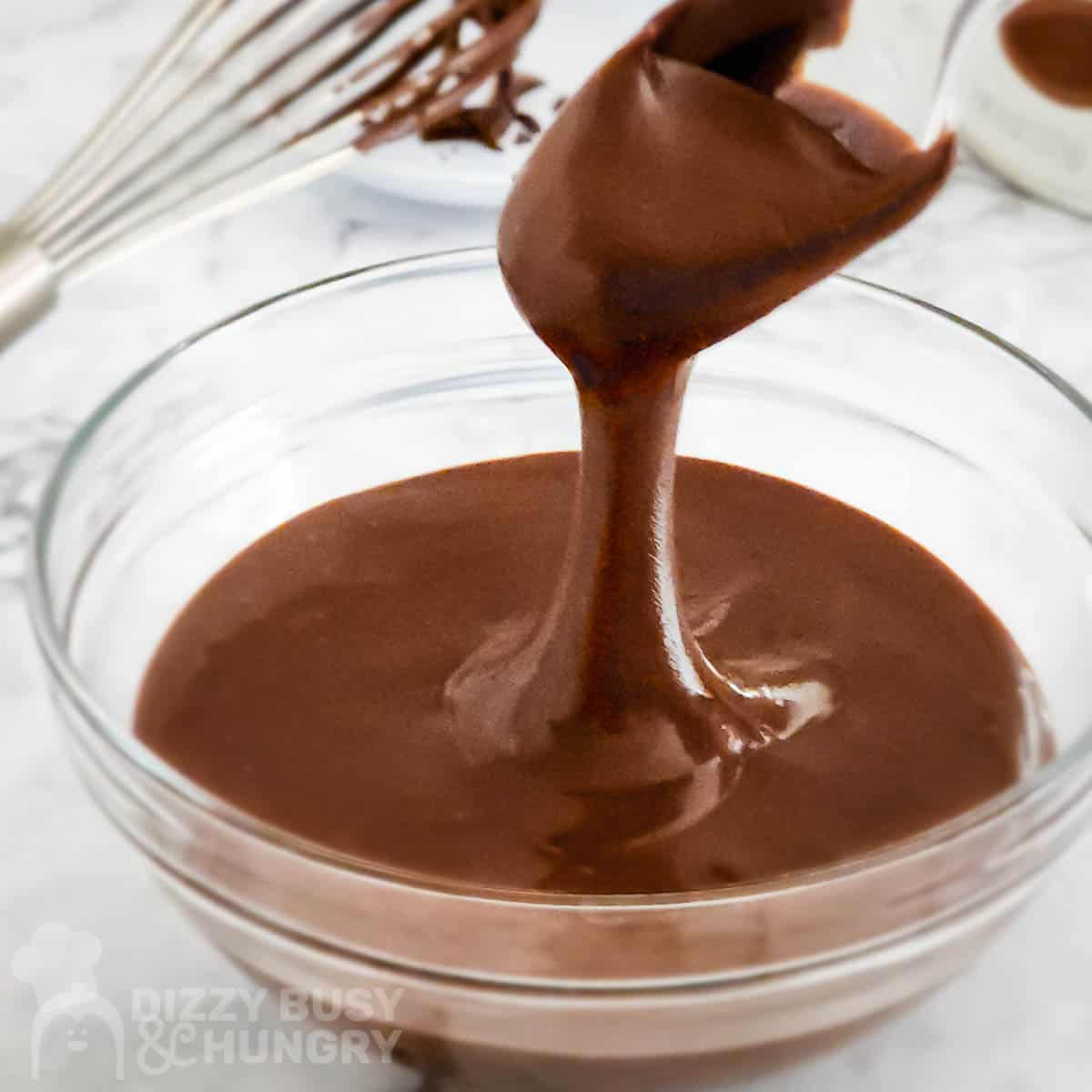Side shot of chocolate glaze being drizzled in a clear bowl full of it with a whisk in the background.