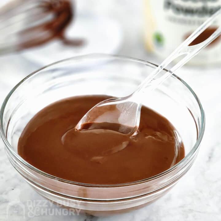 Side shot of a clear spoon scooping chocolate glaze out of a clear bowl.