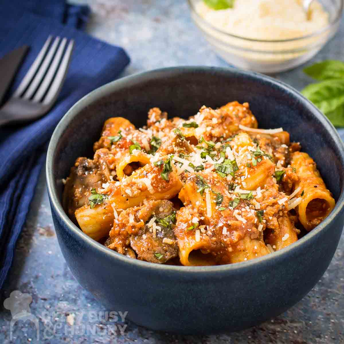 Side shot of rigatoni garnished with cheese and herbs in a blue bowl on a blue speckled surface with a cloth and silverware on the side.