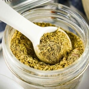 Side shot of poultry seasoning being scooping out of a clear mason jar with a white ceramic spoon.