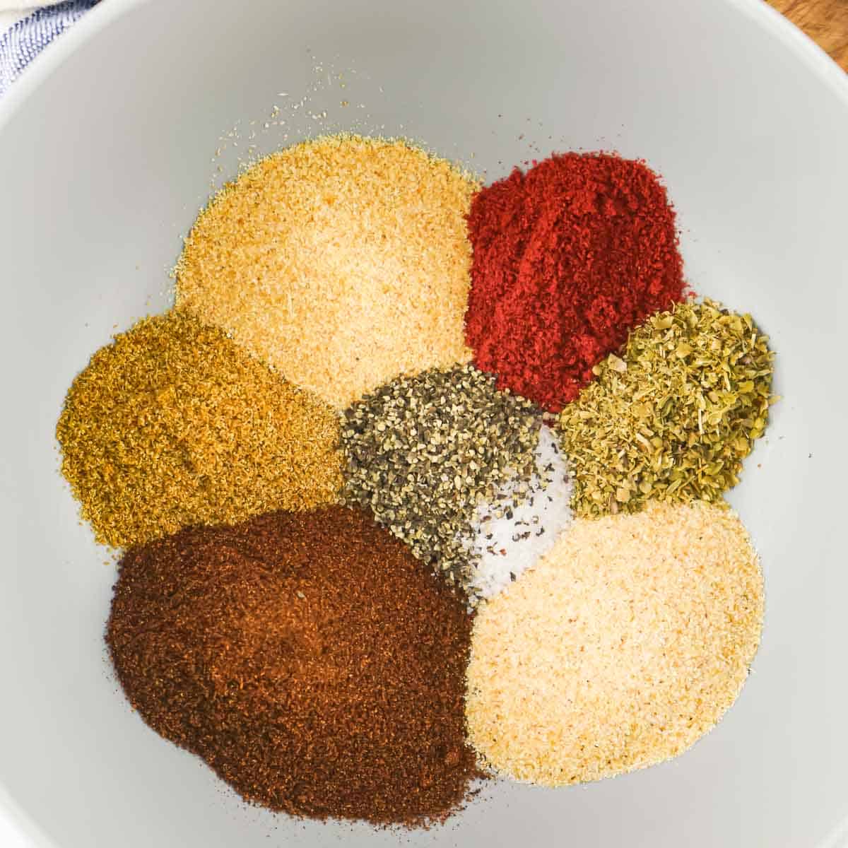 Piles of colorful seasoning arranged in a circle with yellow, red, green, brown, and tan along with salt and pepper in the middle in a white bowl.
