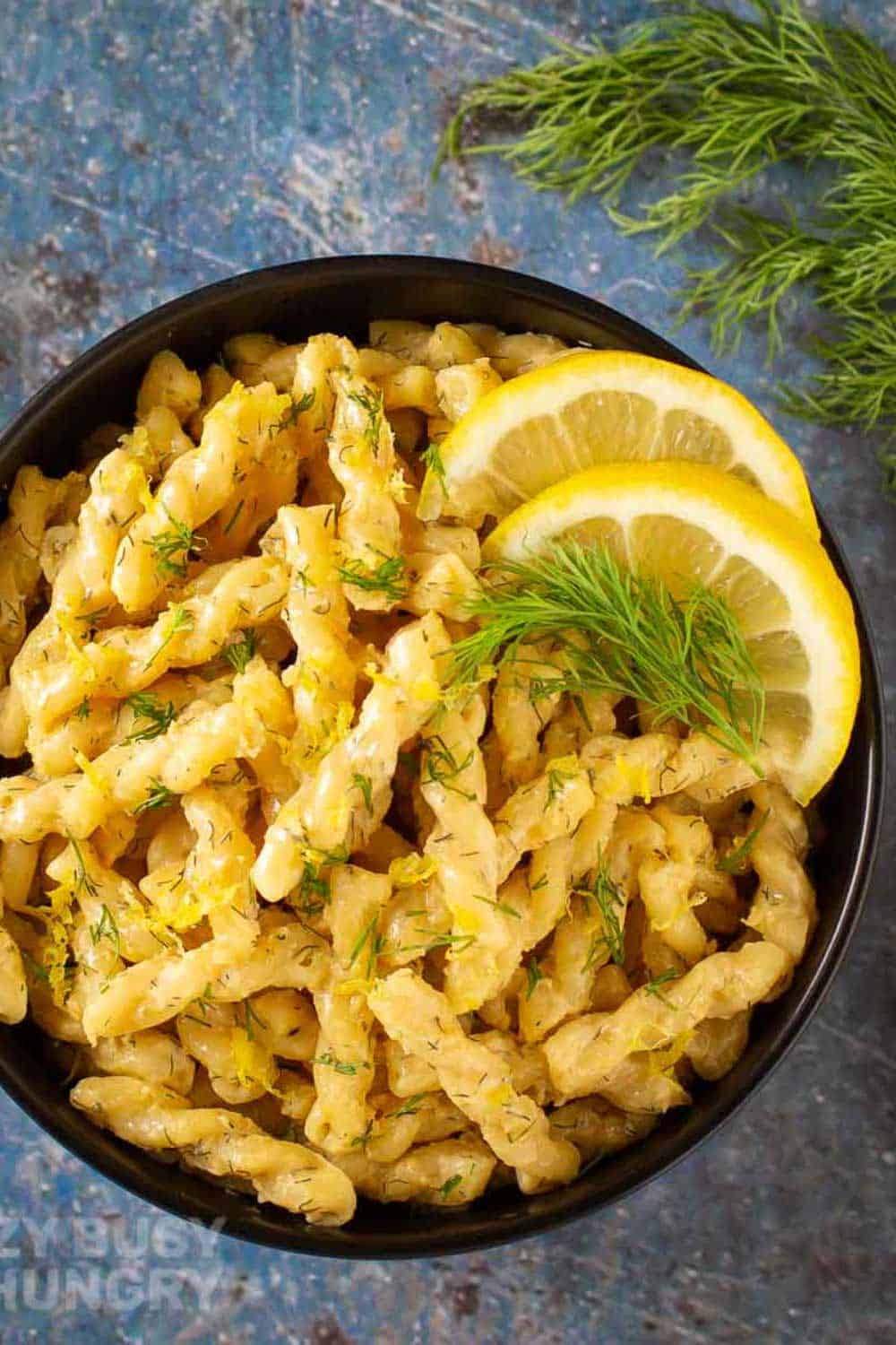 Overhead shot of pasta garnished with lemon zest, dill, and lemon slices in a black bowl on a blue surface with a black fork, dill, and a lemon on the side.