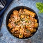 Overhead shot of rigatoni garnished with cheese and herbs in a blue bowl on a blue speckled surface with a blue cloth and basil on the side.