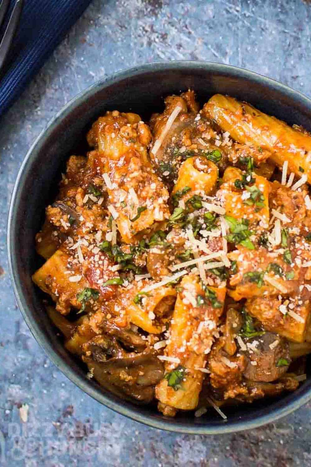 Overhead shot of rigatoni garnished with cheese and herbs in a blue bowl on a blue speckled surface with a blue cloth on the side.