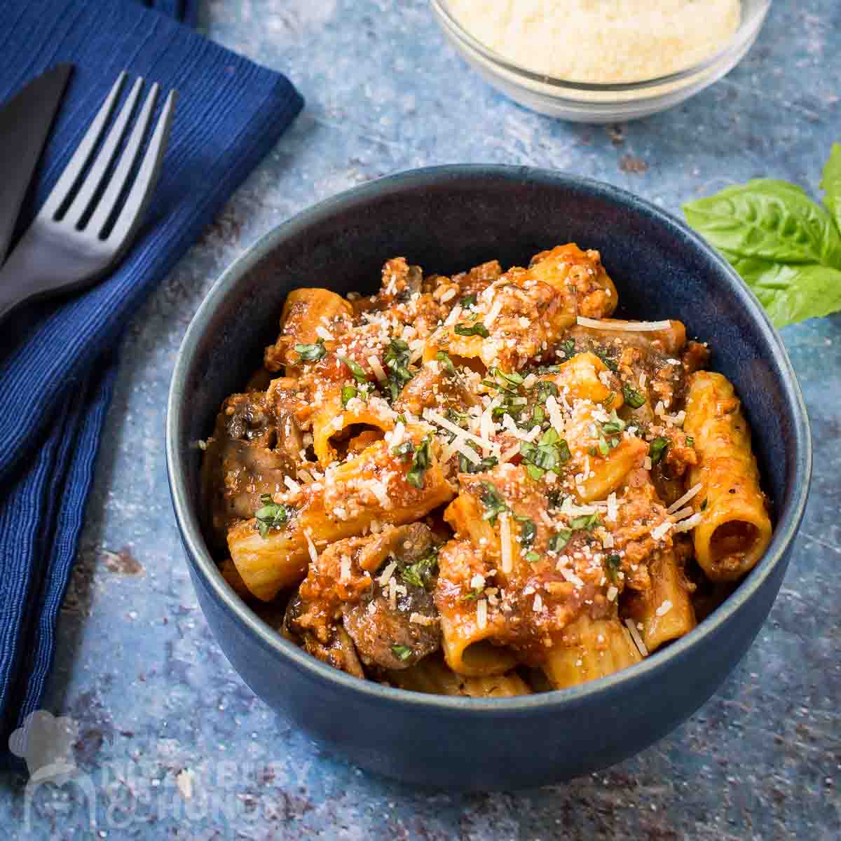 Side shot of rigatoni garnished with cheese and herbs in a blue bowl on a blue speckled surface with a cloth and silverware on the side.