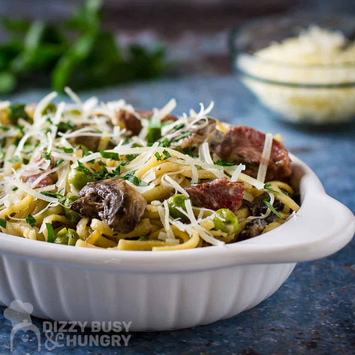 Side shot of turkey tetrazzini garnished with cheese and herbs in a white oval bowl on a speckled blue surface with a bowl of cheese and herbs in the background.