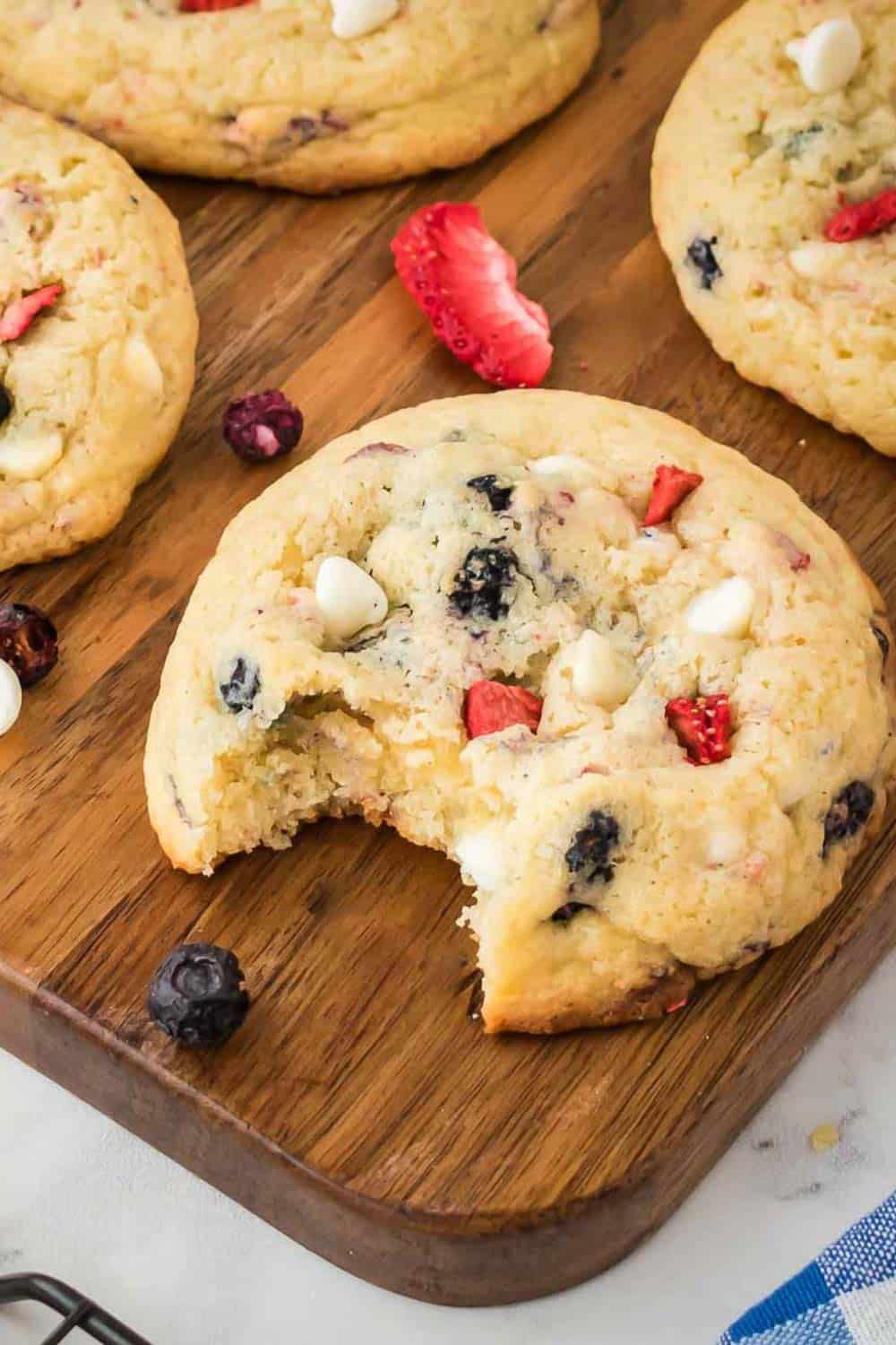 Side shot of multiple cookies, one with a bite taken out, on a wooden cutting board with freeze dried fruits scattered.