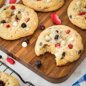 Side shot of multiple cookies, one with a bite taken out, on a wooden cutting board with freeze dried fruits scattered.