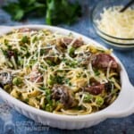 Side shot of turkey tetrazzini garnished with cheese and herbs in a white oval bowl on a speckled blue surface with a bowl of cheese and herbs in the background.