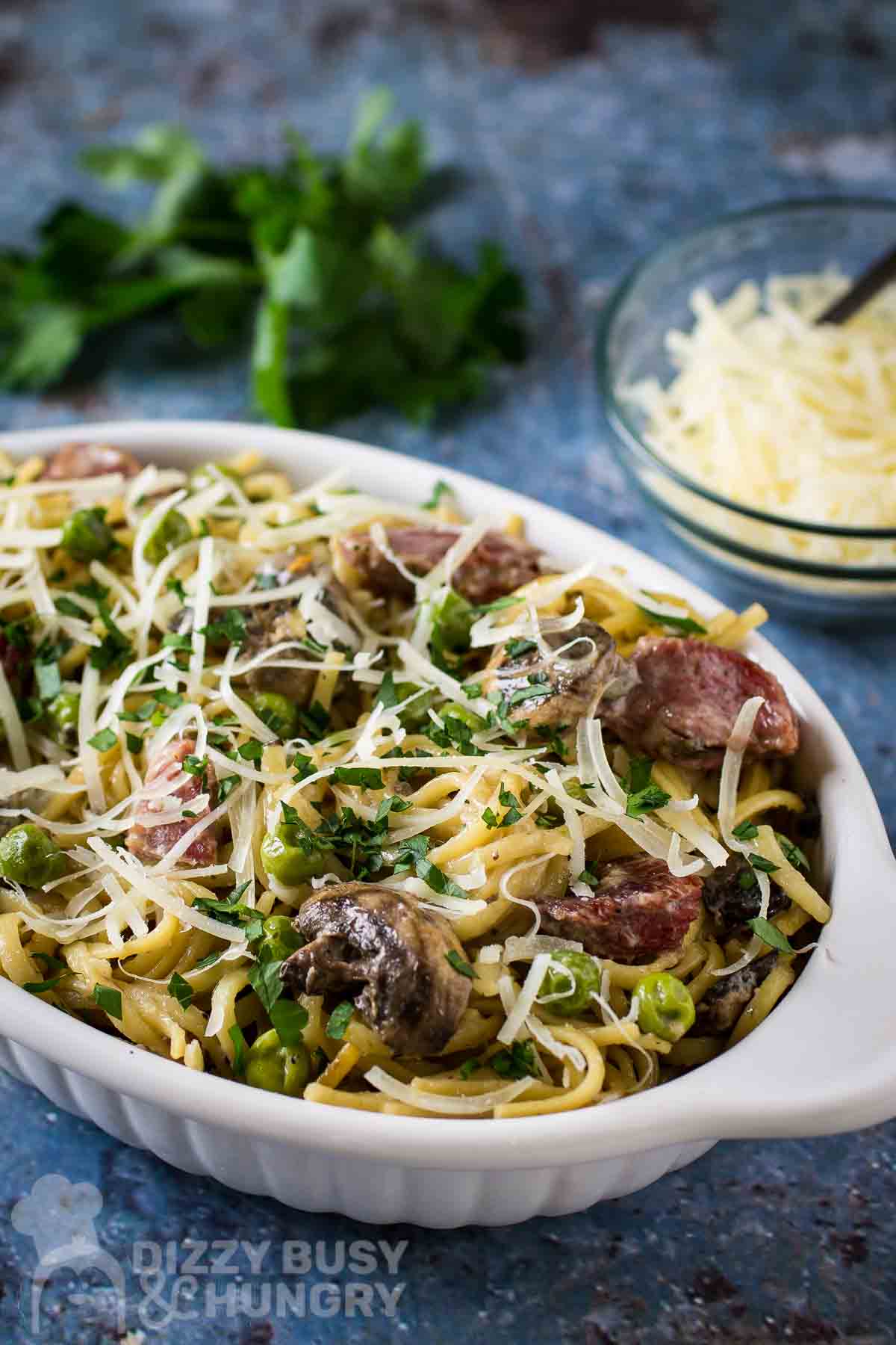 Side shot of turkey tetrazzini garnished with cheese and herbs in a white oval bowl on a speckled blue surface with a bowl of cheese and herbs in the background.