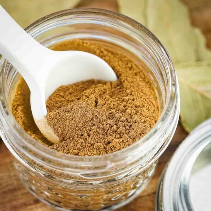 Overhead shot of a white spoon scooping pumpkin pie spice out of a clear mason jar with bay leaves in the background.