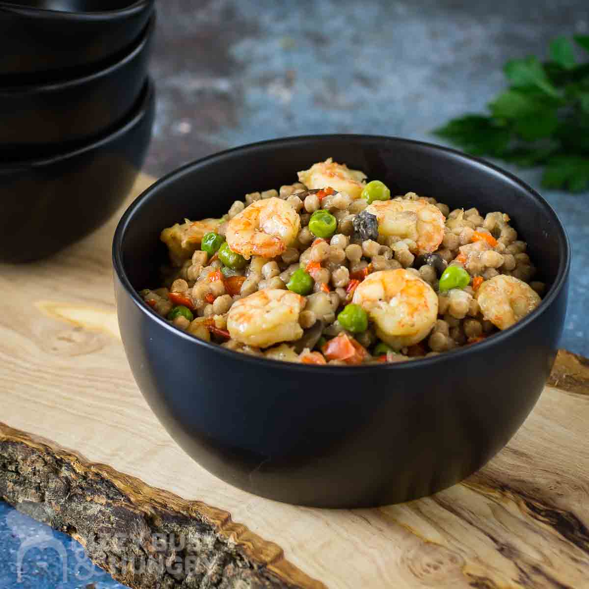 Side shot of shrimp couscous in a black bowl on a wooden cutting board with more black bowls on the side and a blue background.