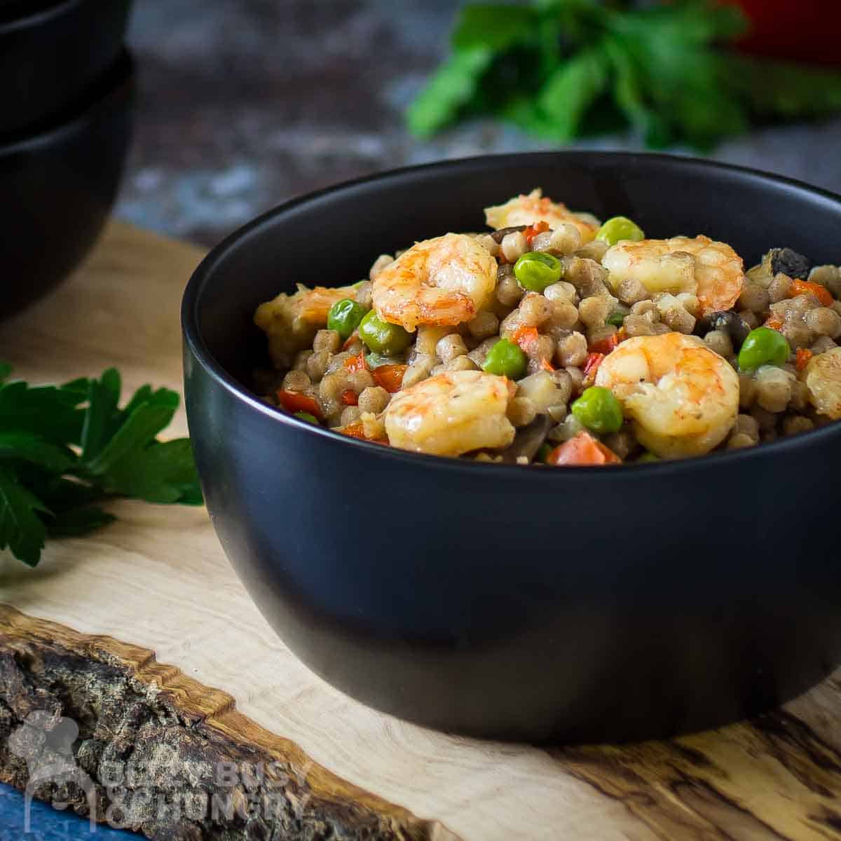 Side shot of shrimp couscous in a black bowl on a wooden cutting board with more black bowls on the side and a blue background.