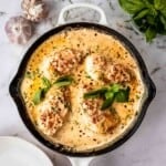 Overhead shot of chicken with sauce in a large skillet garnished with basil on a marble surface with cloves of garlic and basil on the side.