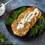 Side angled shot of three salmon patties garnished with dill sauce with a bed of dill on the side on a black plate on a blue marble surface.
