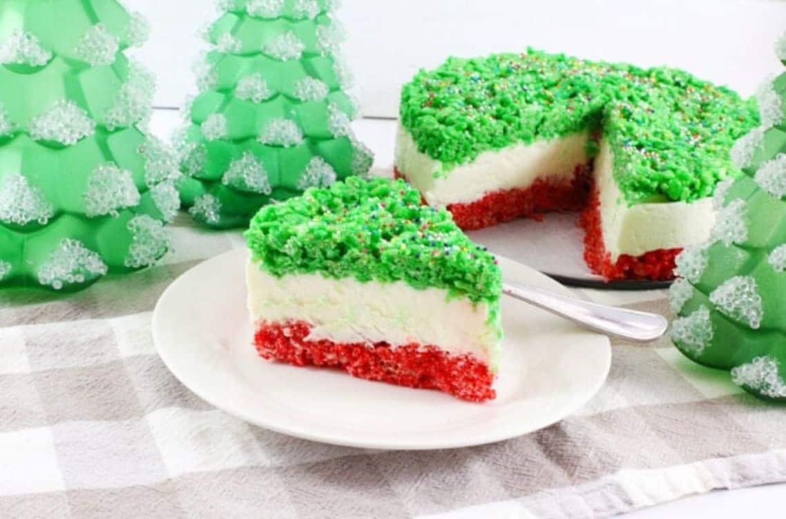 Green, white, and red layered cake with a piece cut out surrounded by green Christmas trees.