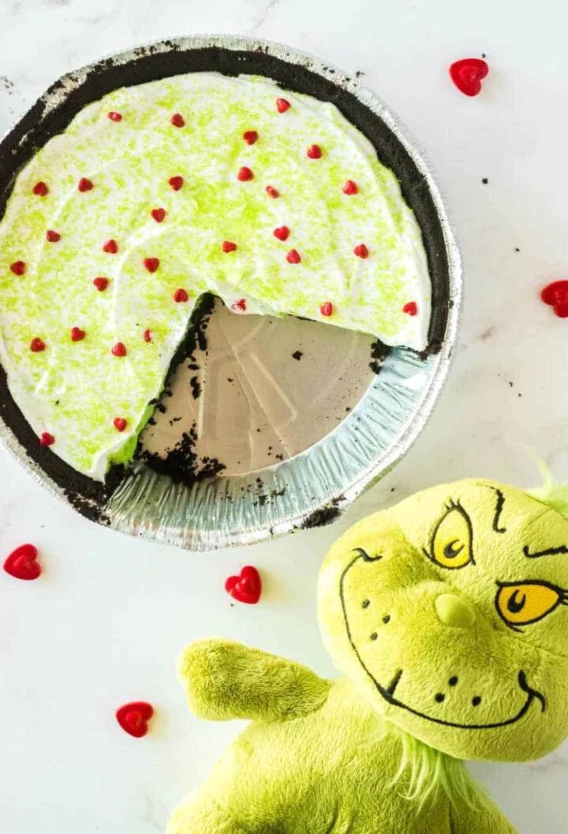 Green pie with chocolate crust and red heart sprinkles next to a Grinch stuffed toy.