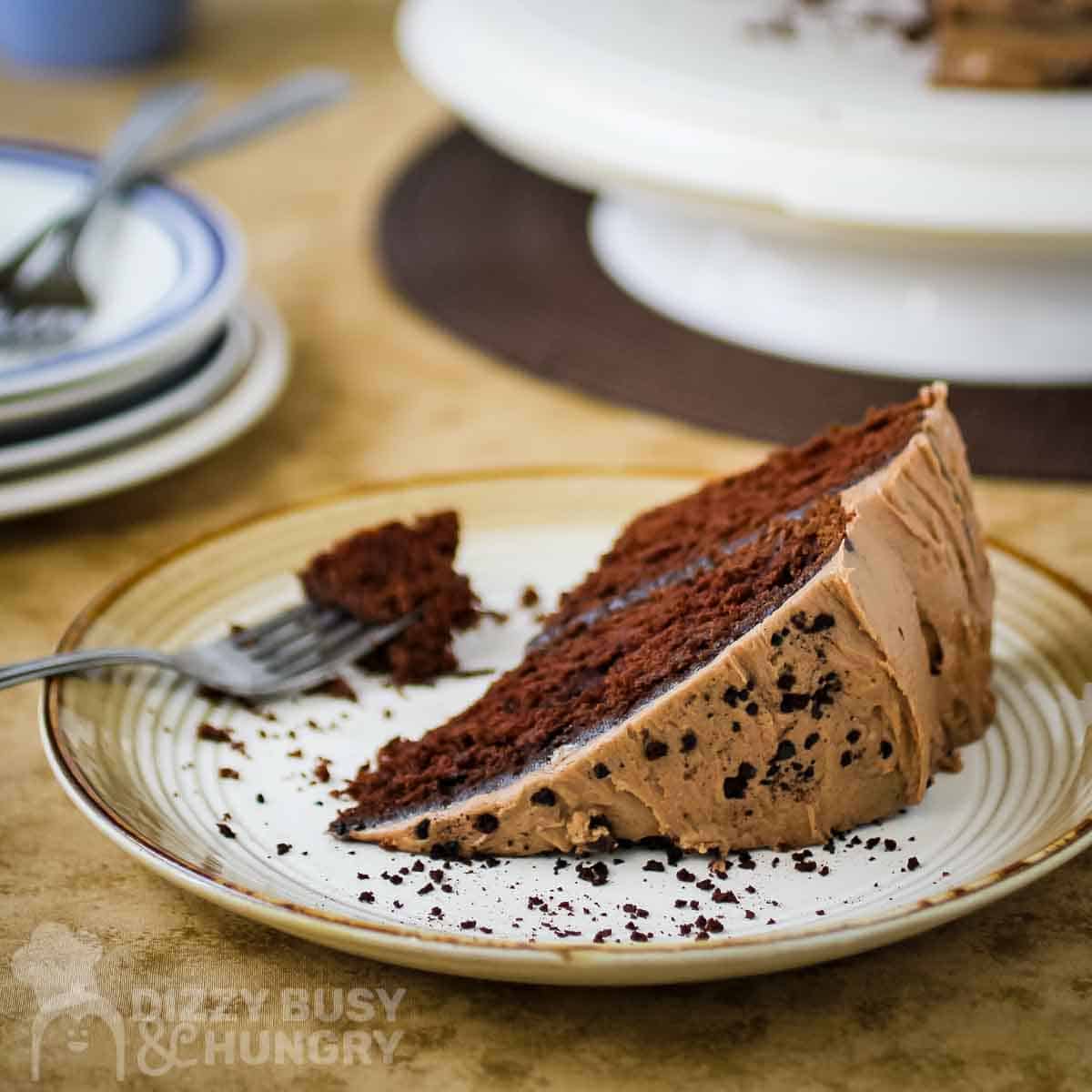 Side view of a slice of hazelnut cake on a white and brown plate with a fork on the side and more plates stacked in the background.