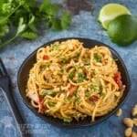Side angled shot of Thai noodles topped with cilantro and chopped peanuts on a black plate on a blue surface with cilantro and lime wedges in the background.