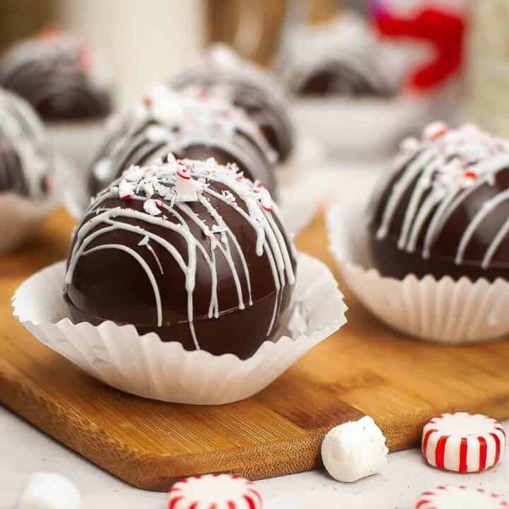 Side shot of mint chocolate bombs in cupcake liners on a wooden cutting board with peppermints scattered around.