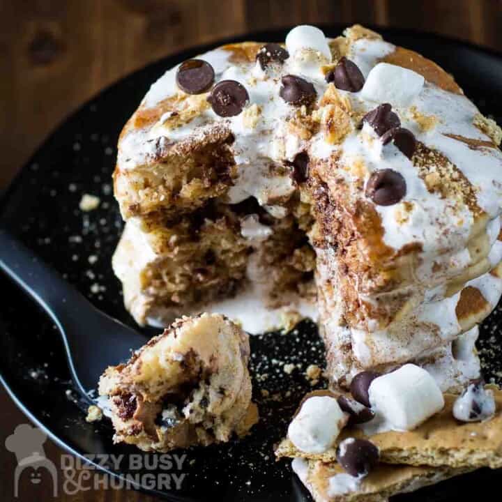 Side close up shot of a stack of smores pancakes with pieces taken out garnished with marshmallow fluff, marshmallows, chocolate chips, cinnamon, and graham crackers on a black plate.