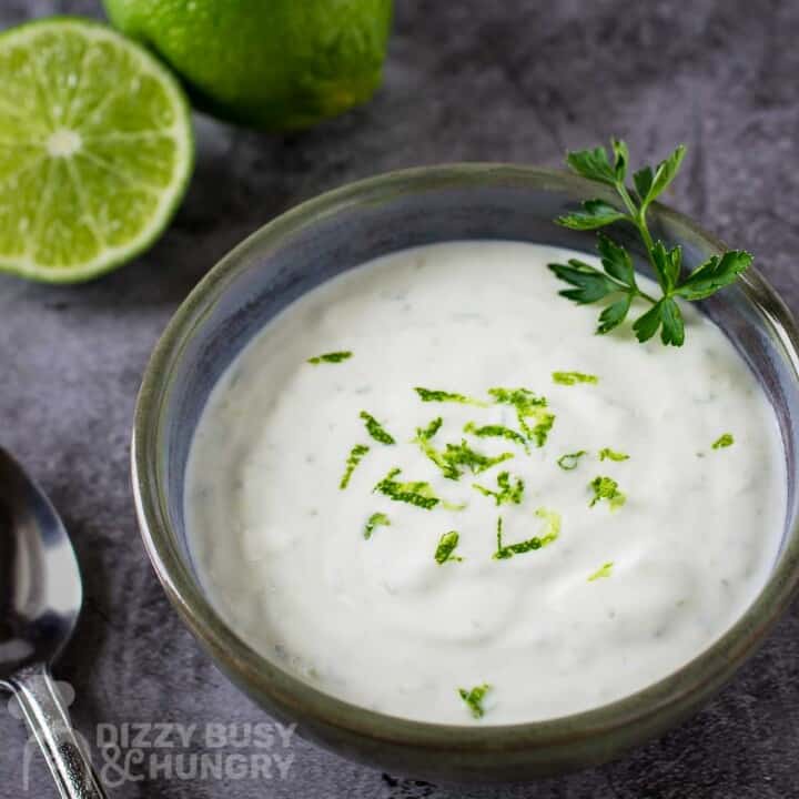 Side angled shot of lime cream garnished with herbs and lime zest in a clear bowl with a spoon and sliced lime on the side.