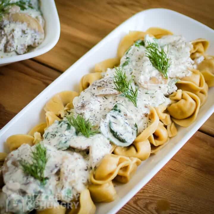Angled shot of mushroom sauce on top of tortellini pasta garnished with dill on a white rectangle plate on a wooden surface.