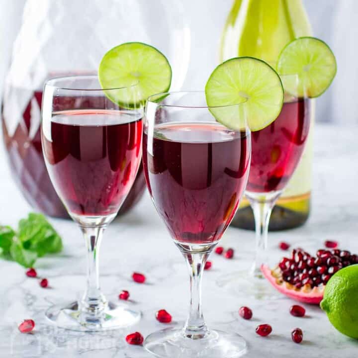 Side shot of three wine glasses with pomegranate cocktail with a lime wedge on the rim, with the pitcher, a wine bottle, and pomegranate arils in the background.