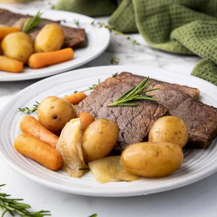 Side shot of rump roast, potatoes, and carrots on a white plate garnished with rosemary with a green cloth in the background.