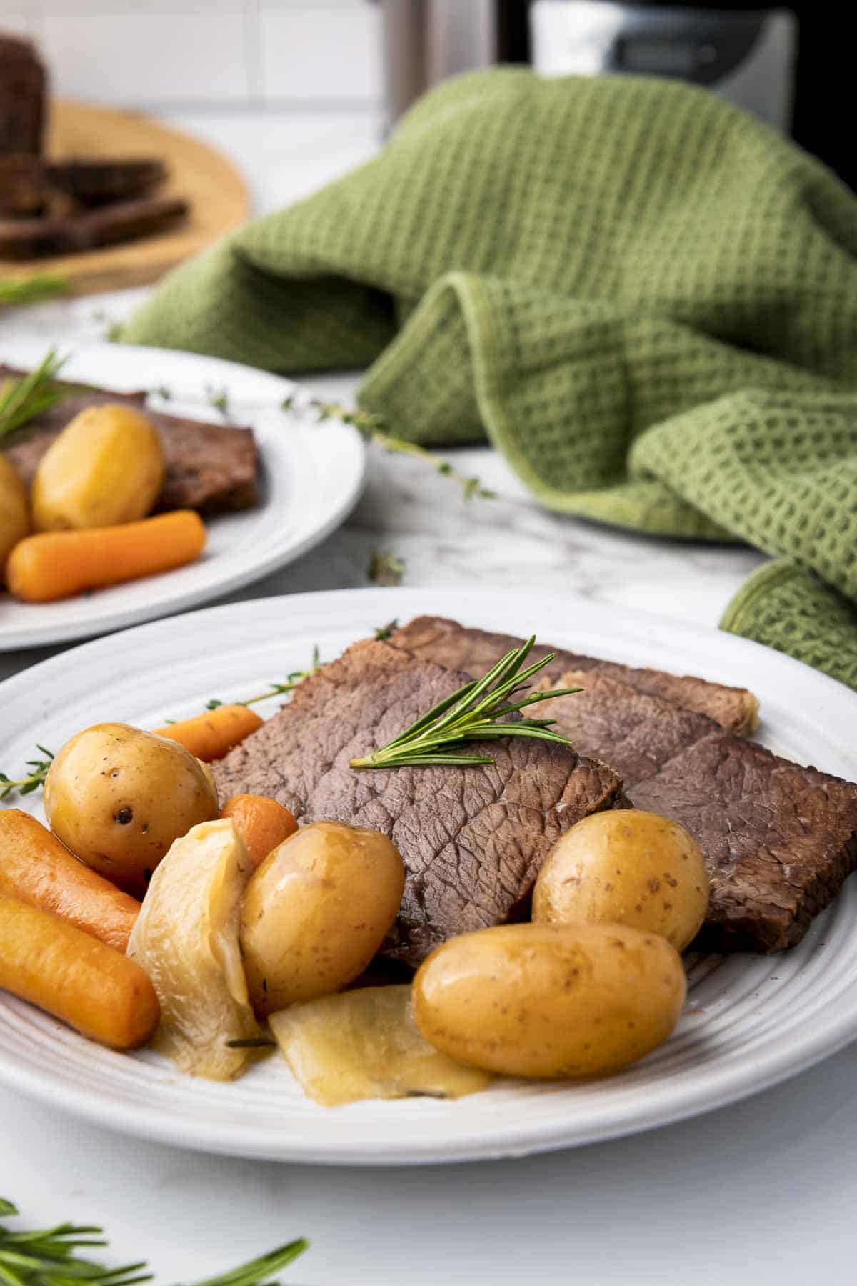 Side shot of rump roast, potatoes, and carrots on a white plate garnished with rosemary with a green cloth in the background.