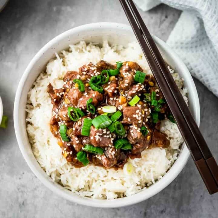 Overhead shot of mongolian beef over a bed of rice garnished with green onions in a white bowl with chopsticks on the side.