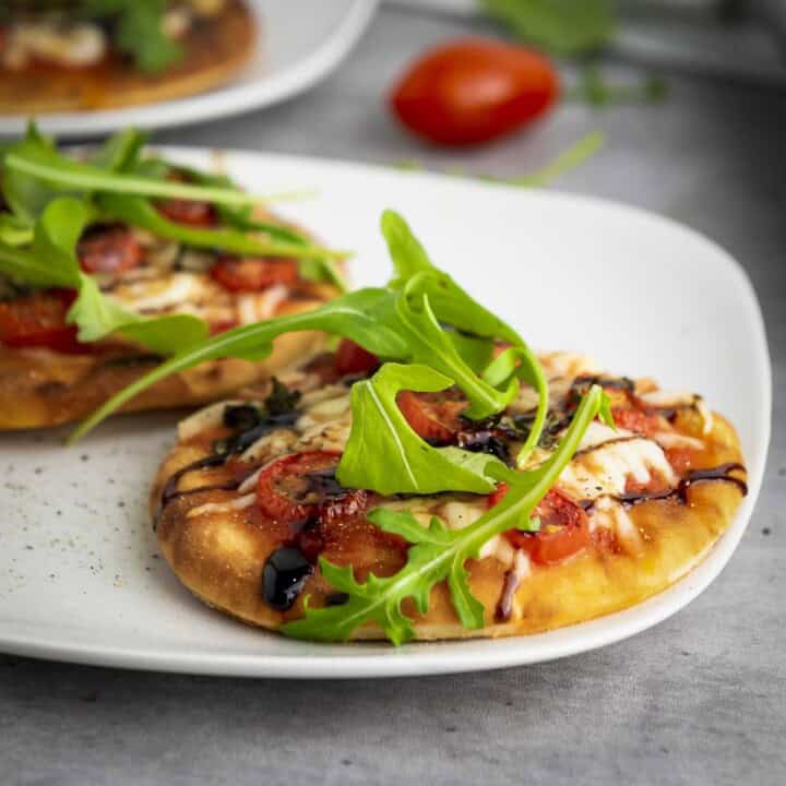 Side shot of two mini caprese pizzas on a white plate with a tomato and greens in the background.