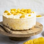 Side shot of the mango cheesecake on a raised wooden cake platter with a knife and mango on the side.