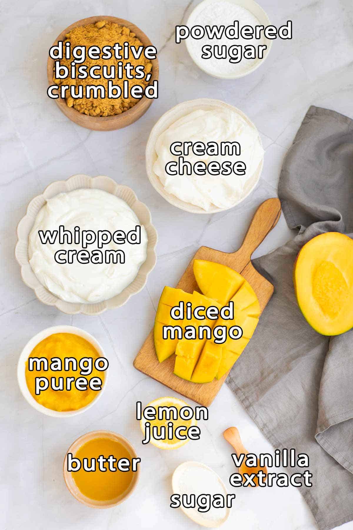Overhead shot of ingredients - digestive biscuits crumbled, powdered sugar, cream cheese, whipped cream, diced mango, mango puree, butter, lemon juice, sugar, and vanilla extract.