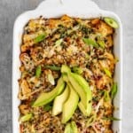 Overhead shot of sushi bake garnished with sliced green onions and avocado in a white ceramic casserole dish on a grey surface.