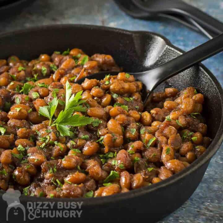 Close up side view of baked beans garnished with fresh herbs in a large skillet with a spatula scooping some out.