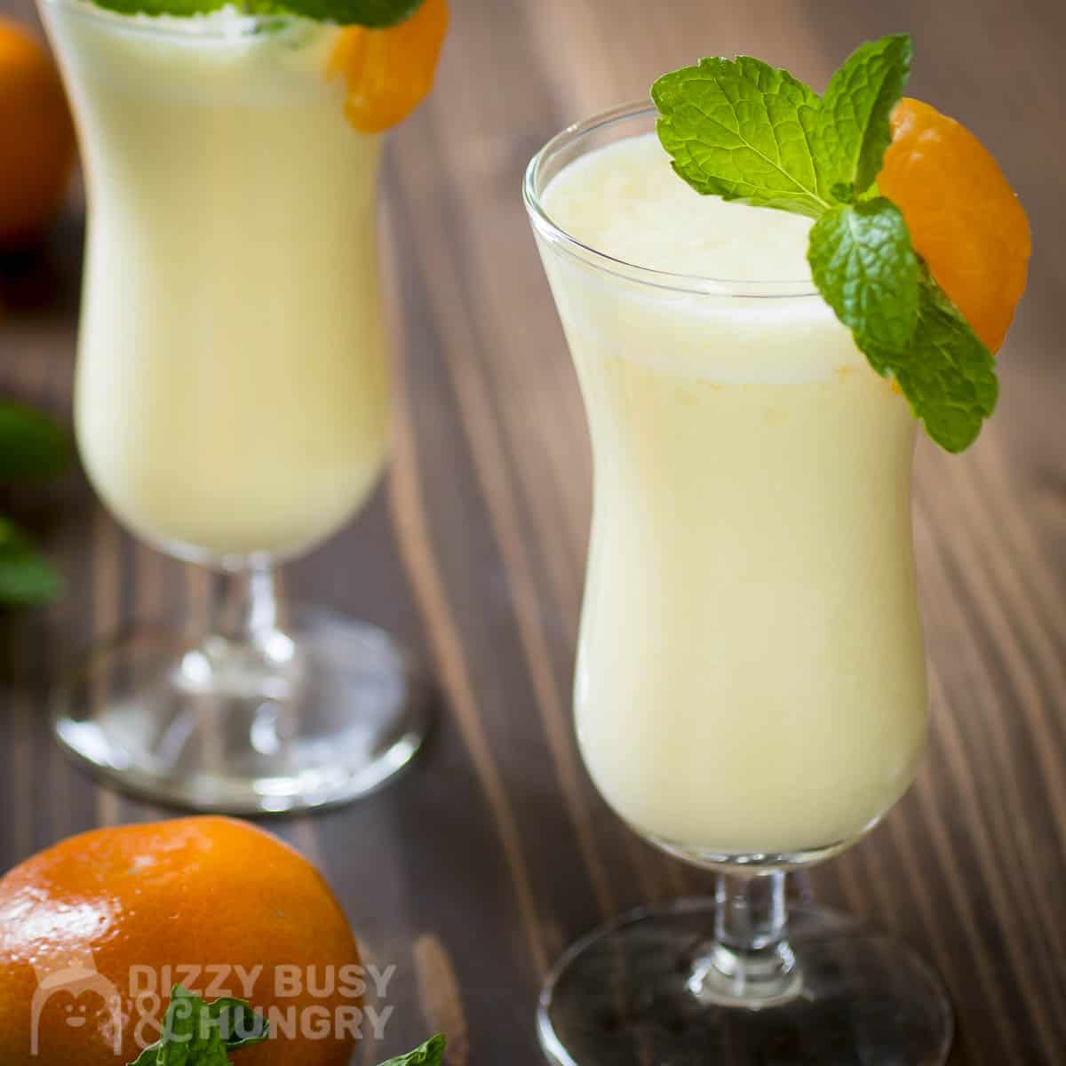 Side shot of two cocktail glasses with orange pineapple smoothie garnished with orange slices and mint with whole oranges and mint sprigs on the side.